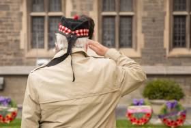 An older man, wearing a ceremonial military cap, salutes a row of wreaths.