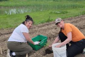 Lisa Owl and Melanie Jeffrey smile as they kneel and place a crookneck squash seedling in the earth.
