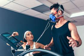 A young man runs on a treadmill, wearing a mask to measure his breathing. A young woman operates the controls.