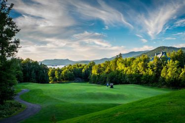 A rolling golf course nestles between sunlit trees, with a hotel, mountains and lake in the background.