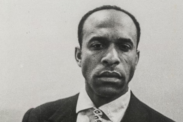 Portrait photography of Frantz Fanon in a suit and tie looking at the camera against a grey background. 