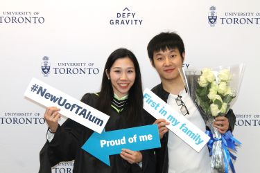 A new grad celebrated graduation at U of T with their friend, holding signs and flowers in front of a “Defy Gravity” backdrop.