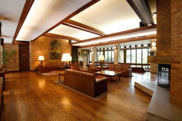 Image of Massey College's Common Room. There is brown furniture placed in the room to invite conversation. There are tables, lamps and a fireplace with large windows. Brick walls and tall white ceiling.