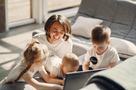 Woman sits on floor next to sofa with three kids looking at a laptop