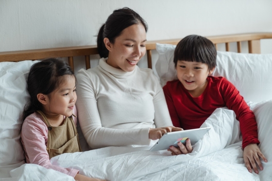 Mother sits with son and daughter on bed looking at tablet