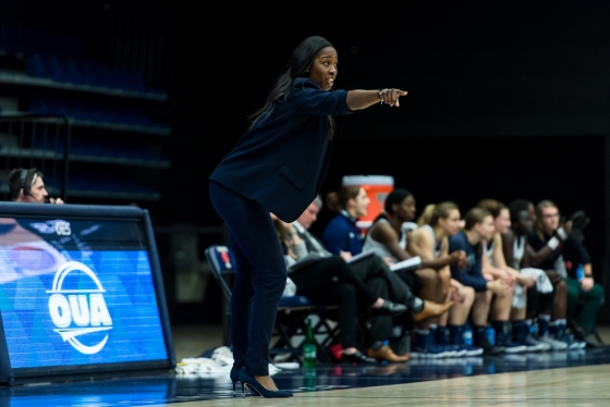 Black woman in navy blue suit stands on edge of basketball court pointing 