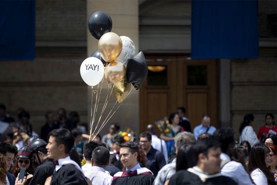 Balloons with the words "YAY!" and "Congrats Grads" catch a shaft of sunlight above a crowd outside Convocation Hall.