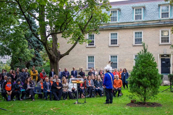 Skahendowaneh Swamp speaks to a crowd gathered on a green lawn. Behind him is a newly planted Eastern white pine tree.
