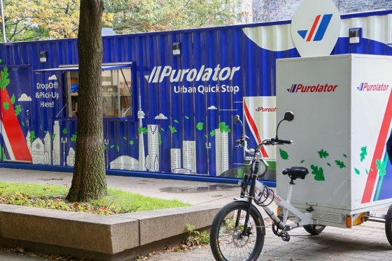 A Purolater delivery bike is parked in front of a shipping container, painted with city skyline and words: urban quick stop.