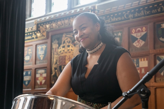 Suzette Vidale smiles happily as she plays a drum in front of a panelled wooden wall at Hart House.