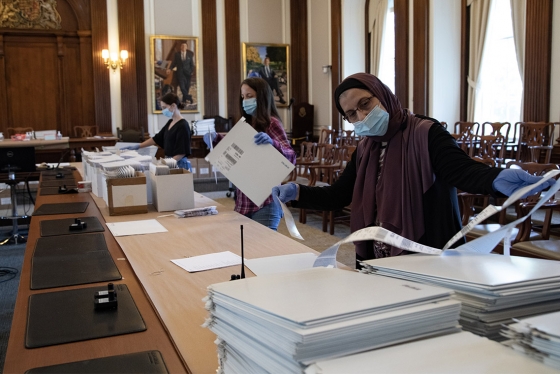 Three women wearing masks and gloves work at a long table loaded with boxes, lists, and piles of envelopes.