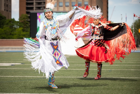 Alanna Pasche and Deanne Hupfield, wearing Indigenous regalia with fringe and embroidery, dance on the turf at Varsity Stadium.