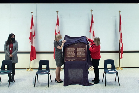At a podium in a classroom, Christine Allen and Christine Loth-Brown lift a cloth to unveil a metal plaque.
