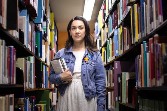 Mikayla Redden stands between tall library shelves, holding a stack of books and looking doubtful.