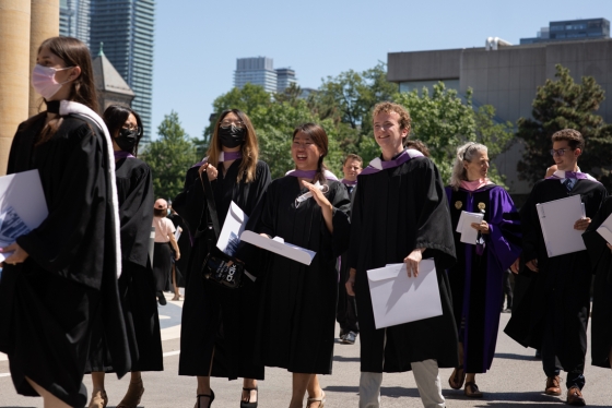A diverse group of graduates, some wearing masks, walk by Convocation Hall, laughing, in their academic robes.