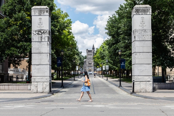 A woman in a mask walks past the U of T gateway on College Street, as the road stretches up to University College behind her.