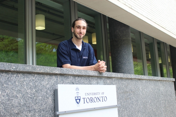 Aidan Gomez smiles, leaning on a stone balustrade above a sign that reads: University of Toronto.