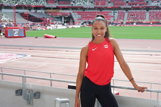 Alice Brown smiles as she stands in the Tokyo 2020 track and field stadium. She wears a shirt with a Canadian flag.