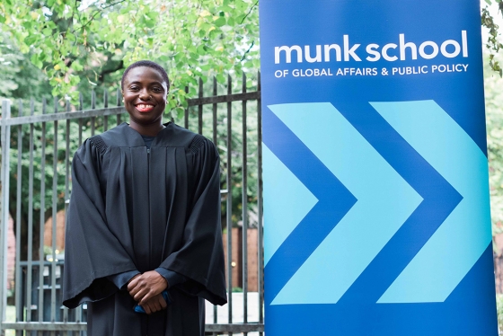 Anowa Quarcoo smiles happily. She is wearing academic robes and standing next to a sign for the Munk School.