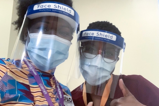 Onye Nnorom and Duate Adegbite, wearing masks and face shields, lean together and give the thumbs up.