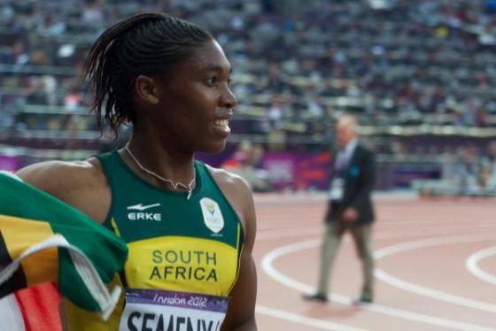 Caster Semenya smiles and waves the South African flag while standing on the track in a stadium.