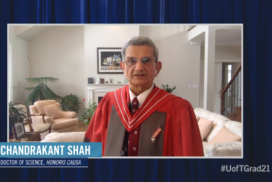 Chandrakant Shah, wearing academic robes, speaks on video from a living room with large plants.