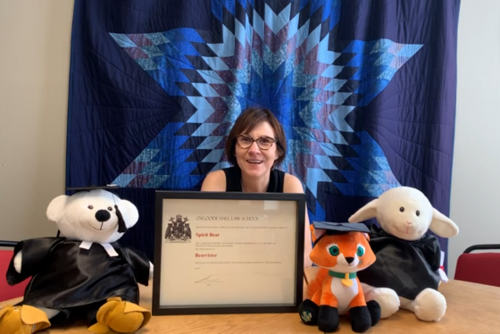 Cindy Blackstock smiles  in front of a quilt with a star pattern. On her desk are stuffed animals in academic robes and hats.