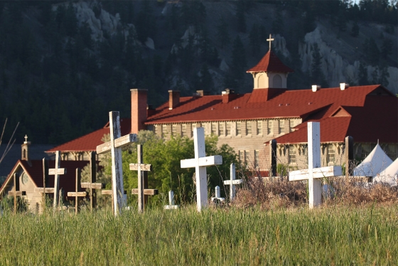 Wooden crosses rise from tall grass. In the background is a large building with a cross on the roof.
