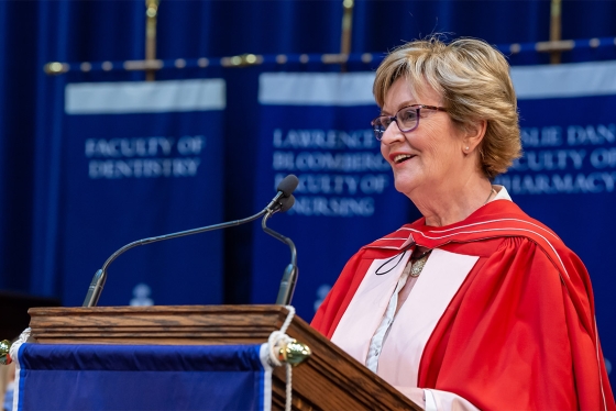 Annette Kennedy smiles as she speaks at the podium in Convocation Hall, wearing academic robes.