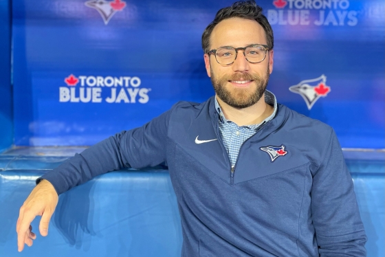 David Lawrence smiles, sitting in the Blue Jays dugout with the team logo behind him.