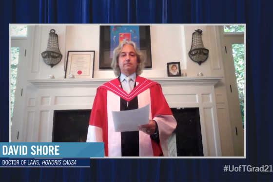 David Shore wears academic robes and speaks in front of a fireplace. On the mantel is his graduation picture as a young man.
