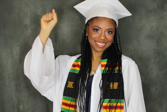 Denessia Blake-Hepburn raises her fist. She is wearing graduation robes and mortarboard hat, and a Black Grad kente scarf.