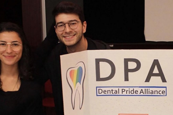 Lucia Santos and Evan Benak smile next to a poster with a rainbow-coloured tooth and the words: Dental Pride Alliance.