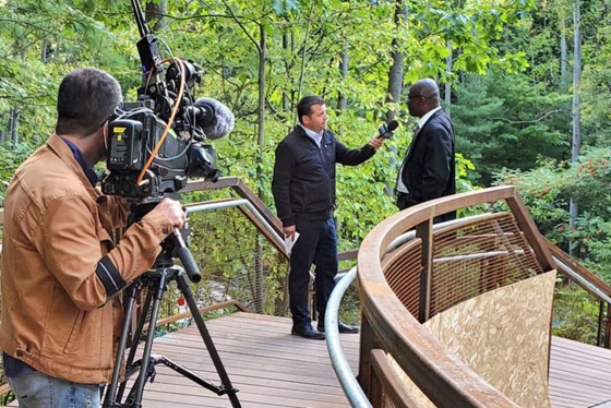 Wisdom Tettey chats with a CTV reporter on a curved wooden bridge, while a man points a large TV video camera at them.