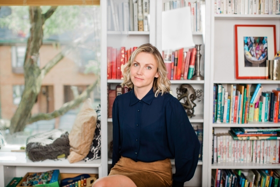 Eva Jurczyk, hands in pockets, sits on a stool in front of bookshelves.