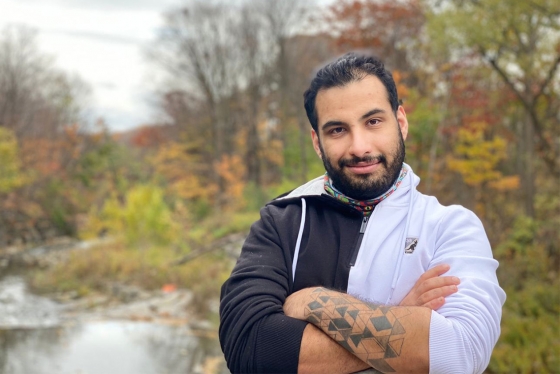 Faraz Khoshbakhtian smiles, standing by a river in a wooded area.