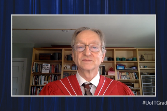 Geoffrey Hinton, wearing academic robes, stands in front of a bookcase and speaks on video.