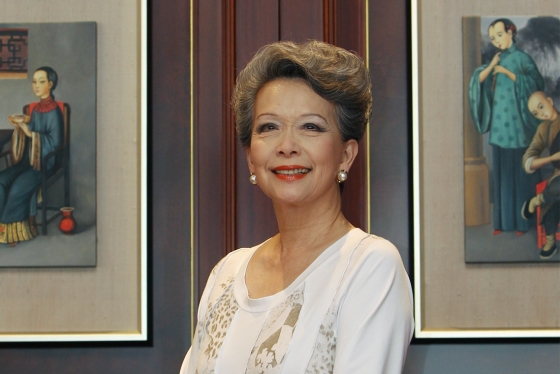 Vivienne Poy smiles, sittingin a room with artwork on the walls showing people in traditional Chinese dress.