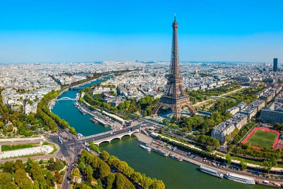 An aerial view of the Eiffel Tower, soaring above tree-lined Parisien streets on the banks of the Seine River.