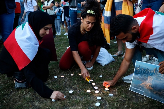 Three people wearing Lebanese flags over their shoulders crouch on the grass and light tea candles.