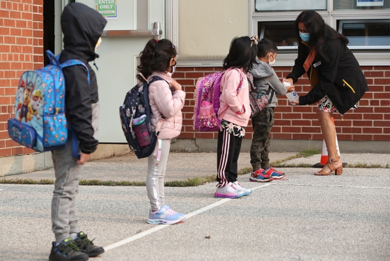 Four small children, wearing masks, stand spaced apart in a playground while a teacher in a mask gives them hand sanitizer.