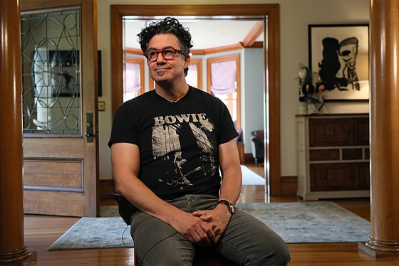 Derek Rossi, wearing a Bowie T-shirt and headphones, smiles as he sits in the hallway of a large house.
