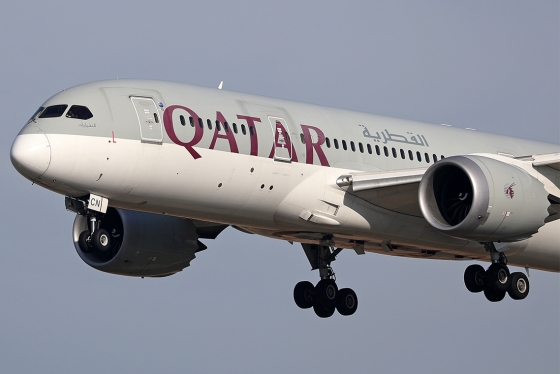 A jumbo jet with wheels down flies low. Letters on the side of the plane read: Qatar.