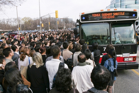 In a pre-pandemic photograph, hundreds of people crowd towards the doors of one lone bus labelled Yonge Subway Shuttle..