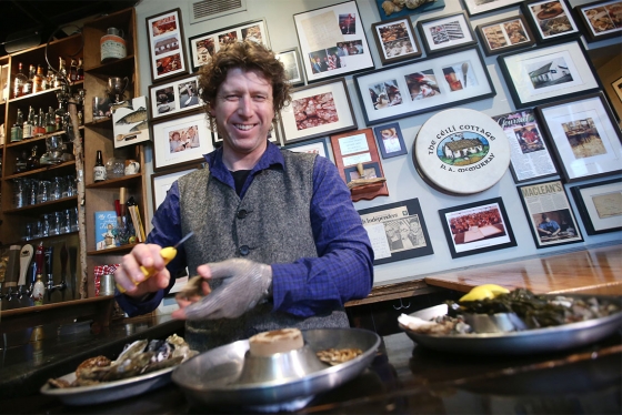 Patrick McMurray smiles as he shucks an oyster at a wooden table near a wall covered with photos and newspaper clippings.