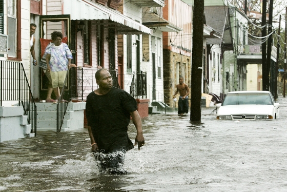 A man wades through knee-deep water down a residential street while people look out their doors.