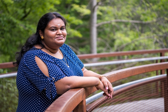 Ilakkiah Chandran smiles, leaning on a railing on a boardwalk in the woods.