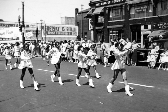 In an undated old photograph, young Black girls in majorette costumes march along a street lined with smiling white people.