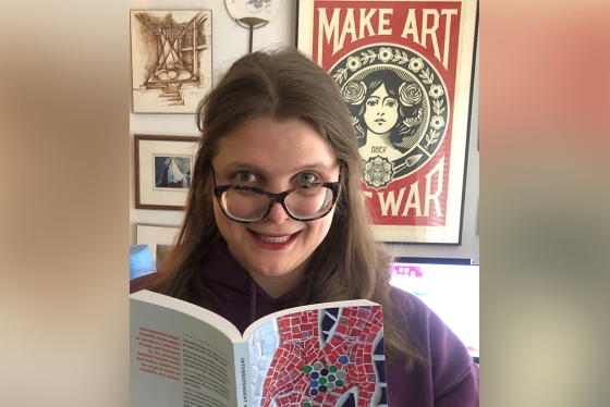 Jessica Watkin holds up an open book. Behind her, a poster that says, make art not war. 