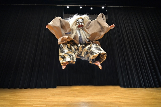 A masked performer jumps up on stage with black drapes behind him